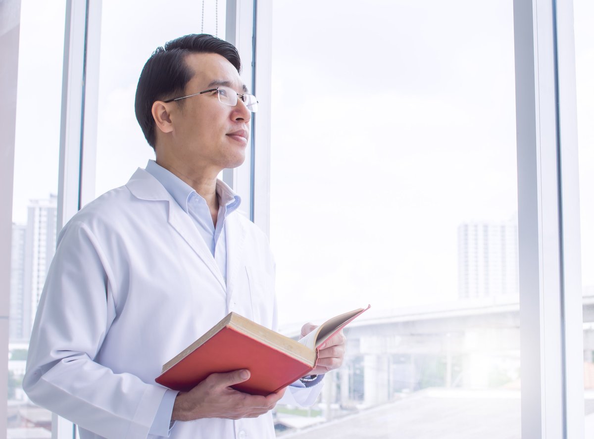 Asian doctor wearing eyeglasses, studying and reading books while standing beside a window