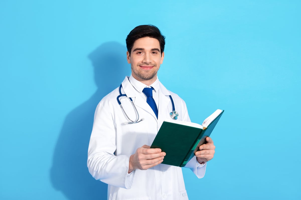 Young male doctor in a white coat and stethoscope holding a book on a blue background